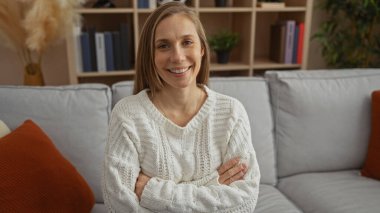 Çapraz kollu bir kadın rahat bir oturma odasında rahat bir koltukta oturur rahat bir şekilde gülümser, rahat bir iç mekan ayarlar, sarışın çekiciliği ve rahat yetişkin tavırları sergiler..