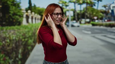 Açık havada, gözlüklü ve kızıl saçlı bir kadın yeşil manzaralı ve kentsel geçmişe sahip şehir sokaklarındaki gürültüden kulaklarını kapatarak güneşli bir günde rahatsızlığını dile getiriyor..