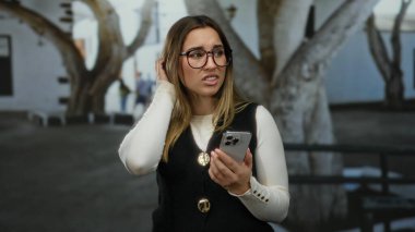 Arka planda ağaçlar, güneşli bir günde siyah yelek ve gözlük takan bir kadın sokakta telefona şaşkın şaşkın bakıyor..