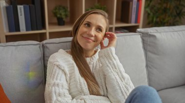 Sıcak bir oturma odasında kanepede oturan, beyaz bir kazak giyen ve gülümseyen, arka planda kitap ve bitkilerle evde sıcak ve davetkar bir atmosfer yaratan bir kadın..