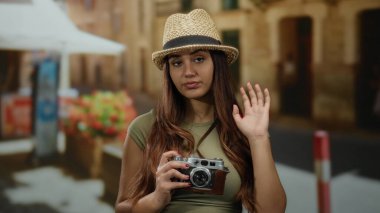 Güneşli caddede kamera dalgaları olan İspanyol bir kadın şapka takıyor. Canlı açık hava alanlarını keşfediyor. Seyahat anlarını yakalıyor.