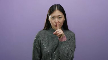 Sessizliği işaret eden bir kadın, parmak dudaklarında, soyutlanmış mor bir arka plan karşısında duruyor, sakin bir tavırla güzelliğini ve Asyalı etnik kökenini vurguluyor..