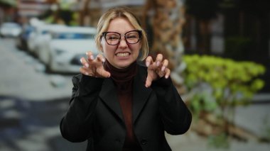 Gözlüklü, gülümseyen bir kadın park etmiş arabaları ve arka planda yeşilliği olan bir şehir caddesinde neşeli pençeler yapıyor..