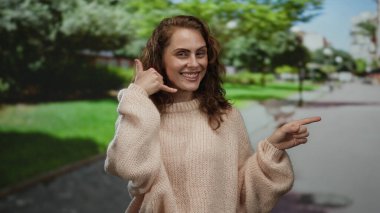 Parkta gülümseyen bir kadın, bir telefon çağrısı sembolü yerleştirirken, güneşli bir gökyüzünün altında yeşil ağaçlarla çevrili gündelik giysiler giyiyor..