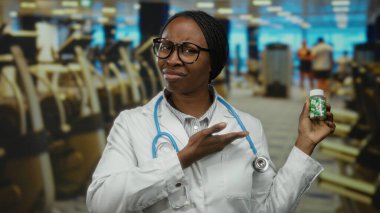 Steteskoplu kadın doktor spor salonunda ilaç şişesi tutuyor, izleyiciye şüpheyle bakıyor, sağlık tercihlerini ve yaşam tarzını tıbbi bağlamda vurguluyor..