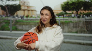 Beyaz kazaklı genç bir kadın şehir manzarası ve ağaçlarla çevrili bir caddede basketbol topunu tutarken keyifli bir gün geçiriyor..
