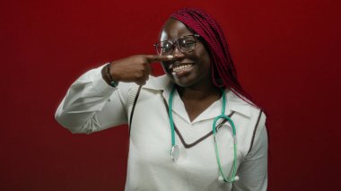 Kırmızı duvara karşı steteskop takan, parmaklarıyla buruna dokunarak gülümseyen Afrikalı Amerikalı kadın, profesyonel sağlık mutluluğunu aktarıyor..