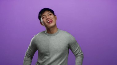 Gri kazaklı genç Çinli adam, izole edilmiş mor arka plan duvarına karşı anlamlı yüz ifadeleri sergiliyor..