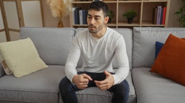 Modern bir apartman dairesinde, kanepede oturan yakışıklı genç adam, sıradan giyinmiş ve düşünceli görünüyor..