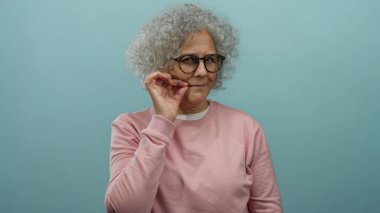 Gri saçlı, gözlüklü ve pembe kazaklı yaşlı kadın mavi arka planda şakacı hareketler yapıyor..