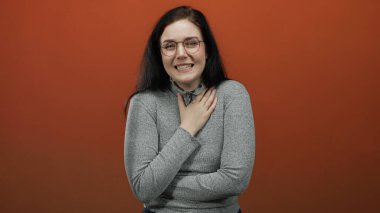 Koyu renk saçlı, gözlüklü ve süveterli bir kadın kırmızı arka plana karşı duygularını ifade ediyor, elini göğsüne koyuyor, rahatsızlık ve şaşkınlık duygularını iletiyor..