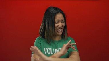 Asyalı bir kadın yeşil gönüllü tişörtüyle canlı kırmızı bir arka plana gülüyor, izole bir ortamda neşe ve toplum desteğini simgeliyor..