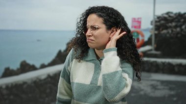 Genç İspanyol kadın, arka planda okyanus ve gökyüzü olan bir deniz kenarında el pençe divan duran bir jest yapıyor. Açık hava sahnesine olan ilgisini ve merakını dile getiriyor..