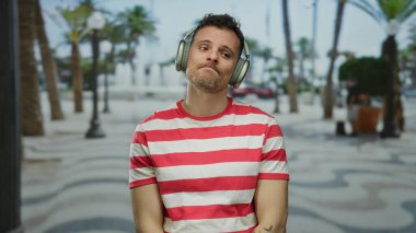 Çizgili tişörtlü genç adam kulaklıkla müzik dinliyor palmiye ağaçları ve bulanık arka planı olan bir şehirde..
