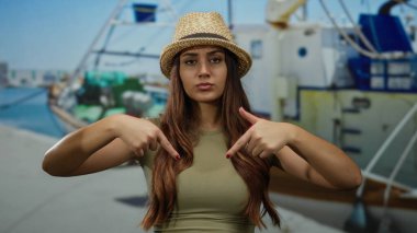 Şapkalı genç bir kadın arka planda teknelerle limanı işaret ediyor açık mavi gökyüzünün altında canlı bir deniz manzarası sergiliyor kendine güvenen bir ifadeyle..