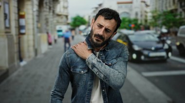 Meşgul bir sokakta kot ceket giyen İspanyol bir adam, dışarıda yansıyor, muhtemelen şehir trafiğinin ortasında omuz ağrısı çekiyor..