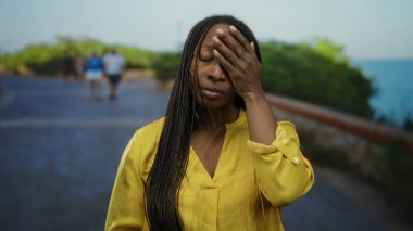 Kıvırcık saçlı, sarı gömlekli, deniz kenarında gezinen bir kadın. Yüzü açık, açık gökyüzünün altında, geçmişi bulanık insanlar.