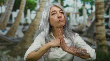 Gri saçlı ve dövmeli bir kadın, içi palmiye ağaçları ve canlı yeşilliklerle dolu bir parkta el hareketleriyle duygularını ifade ediyor..