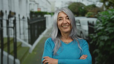 Yeşil ve araçlarla dolu bir sokakta sırıtan gri saçlı yaşlı bir kadın, yeşil bir kazak giyiyor, açık havada mutluluk ve sükunet yayıyor..