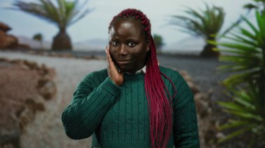 Yeşil kazaklı ve pembe örgülü bir kadın güneşli bir parkta dikiliyor ağaçlarla çevrili, açık havada tefekkür ediyor..