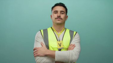 Yansıtıcı yelek giyen İspanyol adam, izole edilmiş yeşil duvara karşı güvenle kollarını kavuşturuyor. Toplum desteği ve dayanıklılığına odaklanmış gönüllü ruhu somutlaştırıyor..