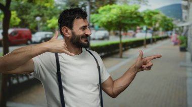 Sakallı İspanyol bir adam, şehir sokağında gülerken gündüz vakti arka planda ağaçlarla arama hareketi yapıyor..