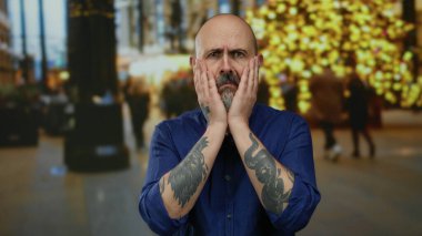 Mavi tişörtlü, sakallı bir adam. Bulanık ışıklarla şehir sokağında endişe ediyor..