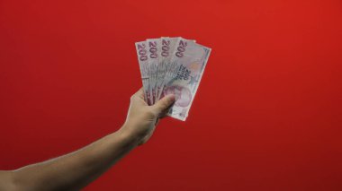 Elinde Türk lirası banknotları tutan adam, izole edilmiş kırmızı bir zemin üzerinde, canlı bir görsel kompozisyonda finans, ekonomi, zenginlik ve döviz alışverişini sembolize ediyor.