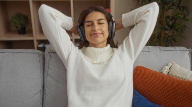 Kanepede oturan, kulaklık takan sıcak bir oturma odasında müziği seven, modern ev hayatı tarzını ve boş zamanlarını sergileyen bir kadın..