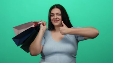 Yeşil arka planda alışveriş torbaları tutan kıvrımlı genç bir kadın alışveriş deneyiminde güven ve neşe gösteriyor..
