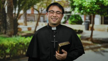 Güneşli parkta İncil 'i tutarken gülümseyen Asyalı rahip, dinsel giysiler giyip, çevrende ağaçlar ve binalar varken huzurlu bir ortamda neşeli bir ifade sergiliyor..