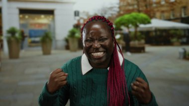 Kalabalık bir caddede yeşil bir kazakla gülümseyen kadın, arka planda canlı renkler ve hareketli bir atmosferi olan şehir ortamında heyecan gösteriyor..