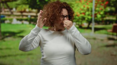 Beyaz gömlekli kadın, parkın dışında, gün boyunca yeşillikle negatif duygularını ifade ediyor. Kıvırcık saçlı ve gözlüklü hoş olmayan bir kokuyu işaret ediyor..