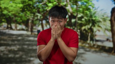 Parkta şok olmuş yüzü olan genç bir adam ağaçların altında kırmızı gömlek giyiyor açık havada güneşli bir arka plan.