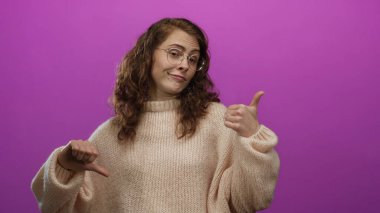 Gözlüklü ve süveterli bir kadın pembe arka planda parmaklarını öne ve arkaya gösteriyor. Canlı bir stüdyoda izole edilmiş neşeli bir ifadeyle karışık duygular ifade ediyor..