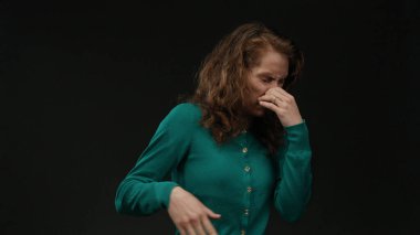 İzole edilmiş siyah arka planda iğrendiğini ifade eden bir kadın, yeşil bir kazak giyiyor, elini burnu örtüyor, güçlü duygular taşıyor, kıvırcık saçlar, esmer, ev ortamında giyiniyor, günlük kıyafetler giyiyor.