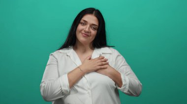 Uzun siyah saçlı, gülümseyen ve ellerini göğsüne koyan beyaz gömlekli bir kadın, izole edilmiş yeşil arka plana karşı duruyor güven, minnettarlık ve pozitiflik taşıyor..