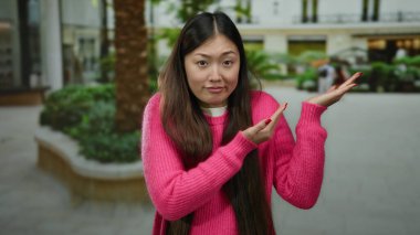 Pembe kazaklı kadın şehir ortamında şaşkın bir ifadeyle dışarıda duruyor. Etrafı yeşillik ve binalarla çevrili. Merakını iletiyor ve sorguluyor..