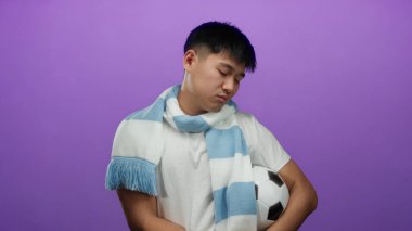 Mor arka planda izole edilmiş futbol topunu tutan mavi eşarplı beyaz tişörtlü genç Çinli adam tefekkür ediyor..