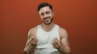 Sakallı genç beyaz adam, izole edilmiş kırmızı arka plan duvarının üzerinden para çekiyor, kolsuz bluz giyiyor, gülümsüyor..