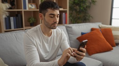 Genç İspanyol bir adam oturma odasında akıllı telefon kullanıyor, rahat bir dekor ve yumuşak ışıklandırmayla çevrili, modern bir apartman dairesinde rahat bir atmosfer yaratıyor..