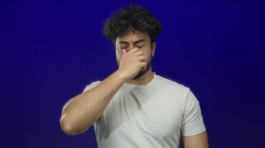 Sakallı genç adam mavi arka planda hapşırıyor, gözlerini kapatıyor ve beyaz gömlek giyerken burnunu tutuyor, rahatsızlığı ve soğuk algınlığı belirtilerini gösteriyor, İspanyol erkek sağlığı.