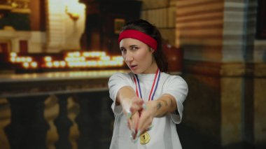 Kilisedeki madalyalı ve saç bandı takan kadın parmaklarını şakaya vurarak atletik ve ruhani temaları mumlarla harmanlıyor..