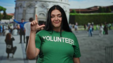 Dışarıdaki kadın kameraya şakacı bir jest yapıyor. Arka planda bir kule var. Gönüllü tişörtü giyiyor..