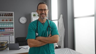 Klinikte önlük giyen İspanyol bir adam tıbbi bir odada kollarını kavuşturarak güvenle duruyor. Hastane işyerinde profesyonellik taslıyor..