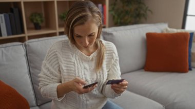 Modern oturma odasında akıllı telefon ve kredi kartı kullanan, rahat koltukta oturmuş şık dekorlarla alışveriş yapan bir kadın..
