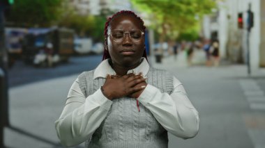 Şehir caddesinde duran bir kadın boğazı ağrıyormuş gibi görünüyor. Elleri boynuna ve göğsüne değiyor, gözlük takıyor ve canlı yeşil ağaç tepe örtüsünün altında gri bir süveter giyiyor..