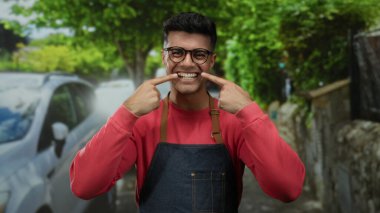 Dişlerine işaret eden gülümseyen gözlüklü ve önlüklü genç adam arka planda ağaçlar ve arabalarla dolu canlı bir şehir caddesinde duruyor..