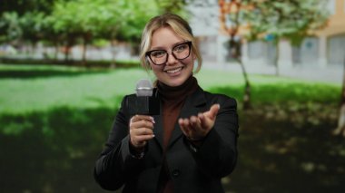 Parkta bir kadın muhabir elinde mikrofon ve işaret parmağıyla elini gözlük takıp güneşli bir gökyüzünün altında yeşillikle çevrili bir şekilde buraya geliyor.