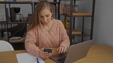 Ofiste çalışan genç bir kadın masa başında otururken dizüstü bilgisayar ve akıllı telefon kullanıyor, görevlere odaklanıyor, iş ve teknoloji ortamını kapalı alanlara yansıtıyor.
